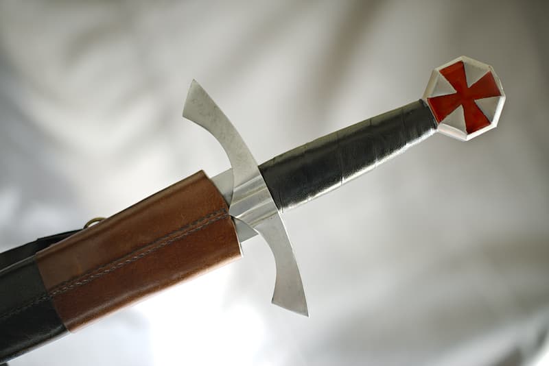 Espada de combate medieval, pomo octogonal con cruz pattée roja, hoja degollada, vaina de cuero marrón y negro con bandolera ajustable