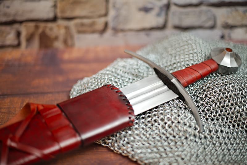Espada medieval forjada siglo XIIᵉ / XIIIᵉ, hoja efecto muelle de doble garganta, vaina de madera y cuero rojo oscuro con cinturón de cuero.
