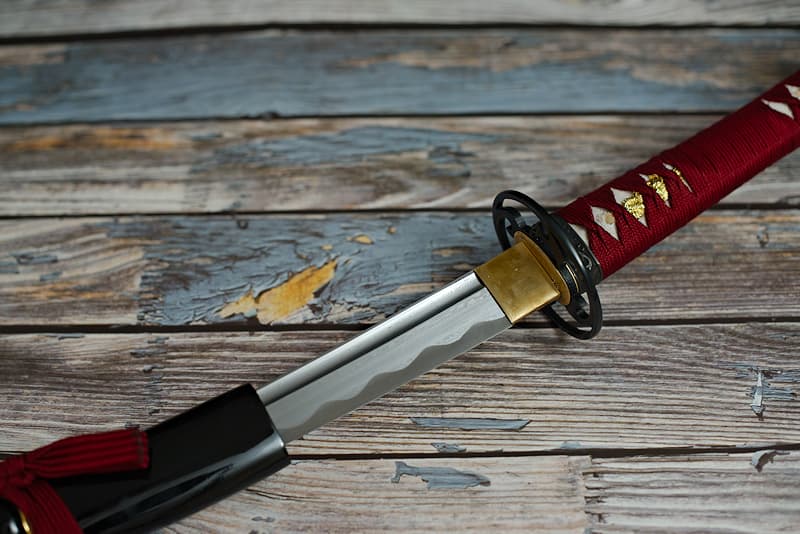 Afilada wakizashi, de forja damasquinada, con vaina negra (saya 鞘), tsuka-ito (柄糸) y sageo rojo oscuro (下緒), entregada en una caja de madera que contenía un kit de mantenimiento y una funda de tela protectora | Nací en un baño de sangre. Renombrado pero discreto, el herrero que me dio a luz fue víctima de su propio éxito. Unos bandidos le asaltaron justo cuando él y mi hermana estaban terminando de forjar la hoja de la katana. No tenían montura, así que no me prestaron atención en el puesto de pulido. No fue de su agrado, porque cuando el maestro samurái para el que estaba destinado descubrió el crimen, encontró un nuevo artesano para crear mi montura de color rojo sangre.