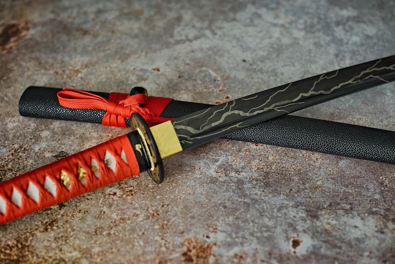 Afilada katana, hoja negra con dibujo de relámpago, con vaina de madera (saya 鞘) recubierta de piel sintética negra tipo shagreen, sageo (下緒) y tsuka-Ito (柄糸) rojo brillante | "Relámpago de acero", ese era el apodo de mi maestro. También me lo pusieron cuando salí de la forja, aún brillante por las llamas.