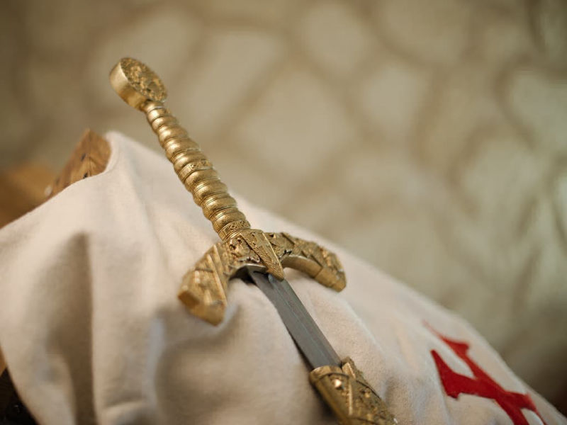 Espada templaria decorativa con vaina roja trenzada, colocada sobre un tabardo templario blanco que lleva el emblema de la cruz roja templaria. Espada templaria decorativa con vaina roja trenzada, colocada sobre un tabardo templario blanco que lleva el emblema de la cruz roja templaria.
