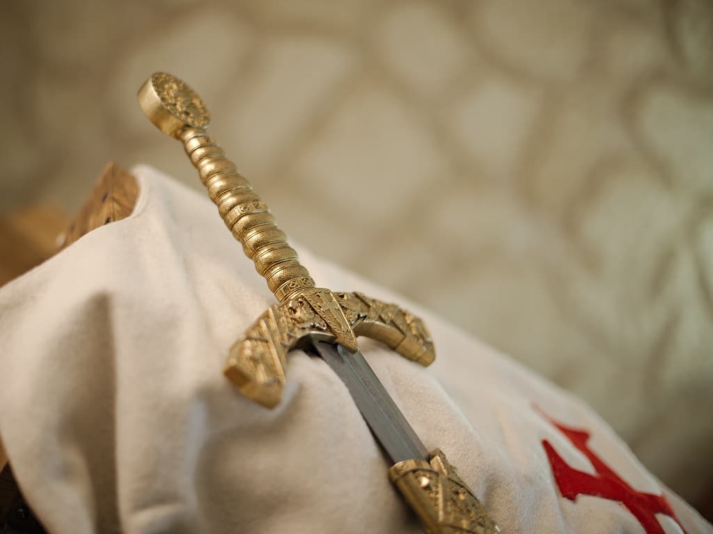 Espada templaria decorativa con vaina roja trenzada, colocada sobre un tabardo templario blanco que lleva el emblema de la cruz roja templaria. Espada templaria decorativa con vaina roja trenzada, colocada sobre un tabardo templario blanco que lleva el emblema de la cruz roja templaria.