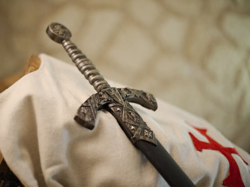 Espada templaria decorativa con vaina negra, colocada sobre un tabardo templario blanco que lleva el emblema de la cruz roja templaria. Espada templaria decorativa con vaina negra, colocada sobre un tabardo templario blanco que lleva el emblema de la cruz roja templaria.