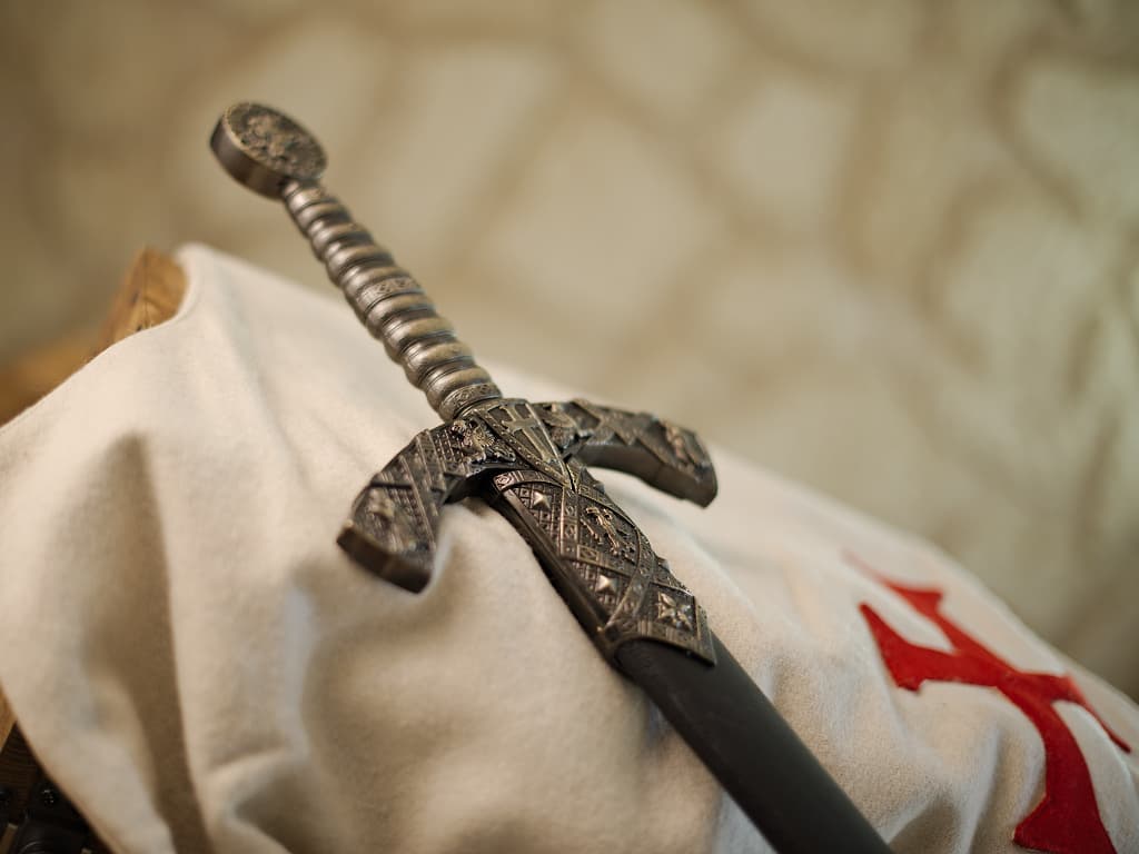 Espada templaria decorativa con vaina negra, colocada sobre un tabardo templario blanco que lleva el emblema de la cruz roja templaria. Espada templaria decorativa con vaina negra, colocada sobre un tabardo templario blanco que lleva el emblema de la cruz roja templaria.