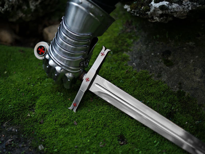 Una mano protegida por un guantelete medieval se apodera de la espada templaria decorativa. Una mano protegida por un guantelete medieval se apodera de la espada templaria decorativa.