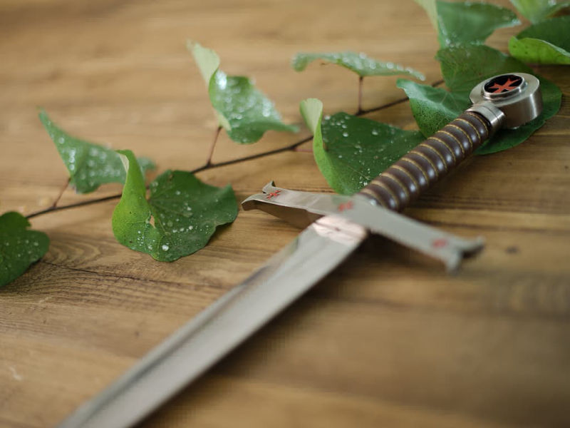 Espada templaria decorativa colocada sobre una mesa medieval de madera, con una rama cuyas ramas albergan gotas de rocío. Espada templaria decorativa colocada sobre una mesa medieval de madera, con una rama cuyas ramas albergan gotas de rocío.
