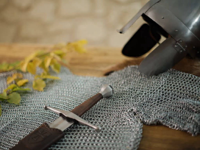 Casco medieval colocado sobre una mesa tredicional, con cota de malla y una espada de asalto medieval.