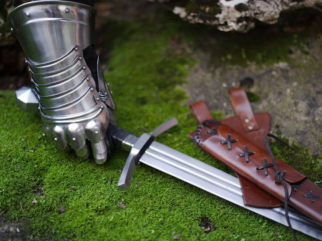La mano de un caballero protegida por un guantelete articulado se apodera de una espada medieval forjada que descansa sobre una roca cubierta de musgo.