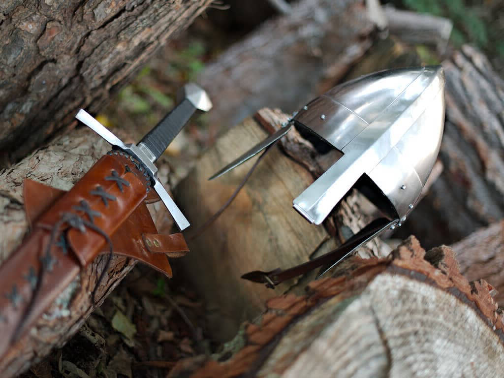 Casque et épée médiévale posés sur des bûches en bois en pleine forêt enchantée Casque et épée médiévale posés sur des bûches en bois en pleine forêt enchantée
