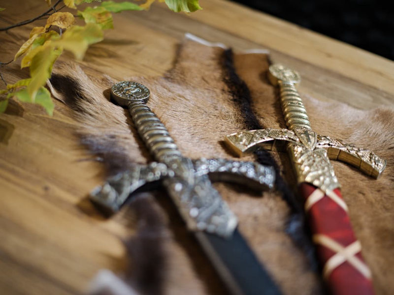 Les deux épées templières de décoration avec fourreau rouge tressé ou noir, posées sur une table médiévale en bois, avec une branche automnale Les deux épées templières de décoration avec fourreau rouge tressé ou noir, posées sur une table médiévale en bois, avec une branche automnale